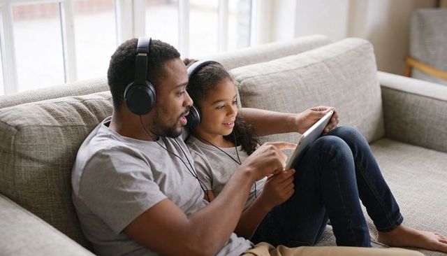 Father and daughter sharing tablet and listening with headphones on cozy living room sofa