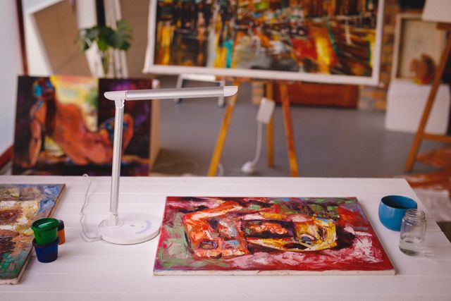 Colorful abstract painting lying on a desk in an art studio. Various art supplies, including paint jars and brushes, are scattered around. Ideal for illustrating creativity, artistic environments, and the process of creating art. Suitable for use in articles about art studios, artist inspiration, and creative workspaces.
