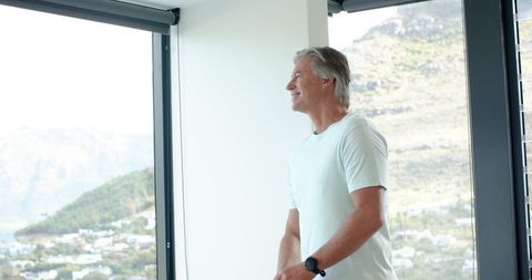 Senior man gazing out large window overlooking hillside from modern home
