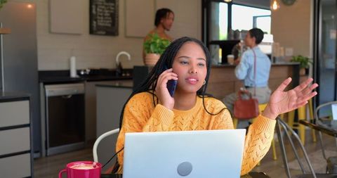 African american woman working remotely in cafe on laptop and phone with coffee