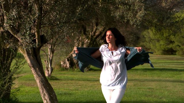 A woman walks in a park enjoying a breezy day while putting on a jacket. Perfect for concepts of freedom, nature, outdoor lifestyles, and capturing peaceful, carefree moments. Good for websites or blogs focused on leisure, outdoor fashion, or mental well-being.