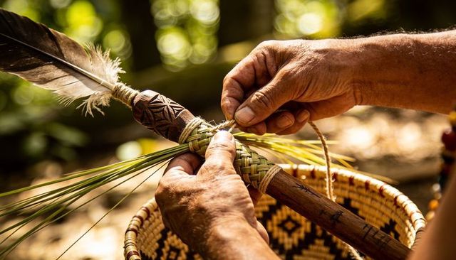 Hispanic artisan weaving green fiber braid around carved wooden staff with feather, basket