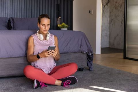 Woman relaxing post-workout wearing leggings and headphones using smartphone in sunlit minimalist be