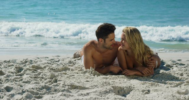This photo shows a young couple lying on a scenic sandy beach with ocean waves in the background, enjoying a romantic moment. Ideal for depicting themes of love, vacations, summer getaways, relaxation, and outdoor activities. Perfect for travel brochures, romantic advertisements, beach-themed promotions, and lifestyle magazines.