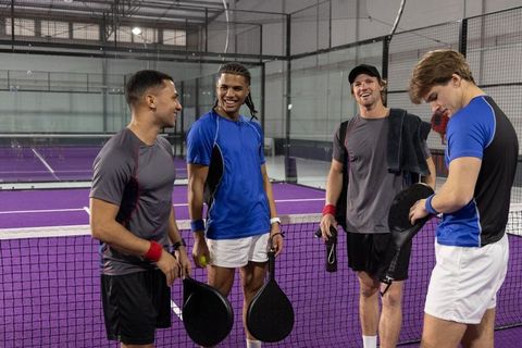 Diverse male teammates chatting and laughing on purple padel court holding paddles and ball
