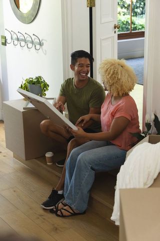 Smiling asian man and african american woman unpacking new home holding artwork in foyer