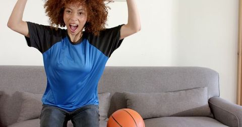 Cheering woman in blue sports shirt celebrating basketball win at home on gray couch