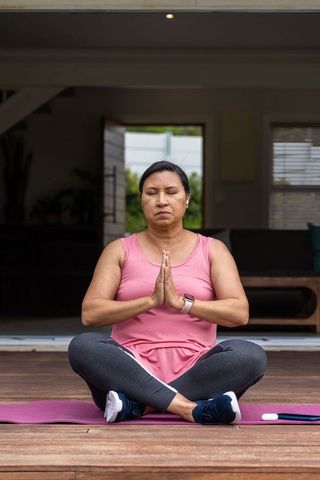 Mature woman meditating on wooden deck doorway on pink mat wearing smartwatch and activewear