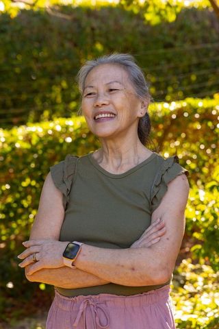 Senior woman standing with arms crossed smiling in sunlit garden wearing smartwatch