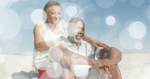 Mature couple smiling and relaxing on sunny beach with bokeh light overlay serene coastal lifestyle