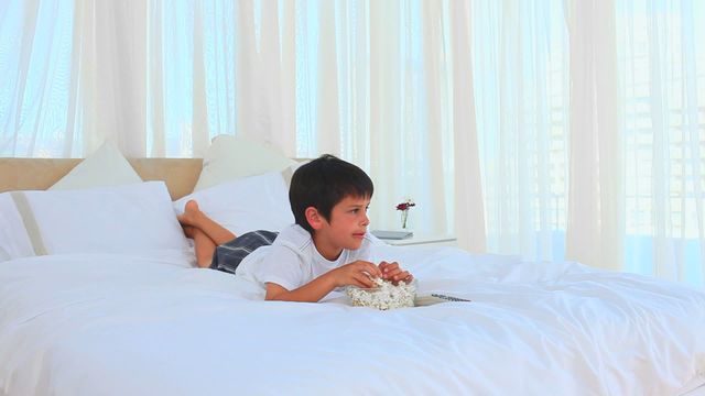 Little boy lies comfortably on a large bed while eating popcorn, with a remote control beside him. The setting is relaxed with soft sheets and plenty of natural light shining through sheer curtains, suggesting a family-friendly and safe home environment. Perfect for depicting moments of leisure, parenthood, or interiors focused on everyday life or family bonding.