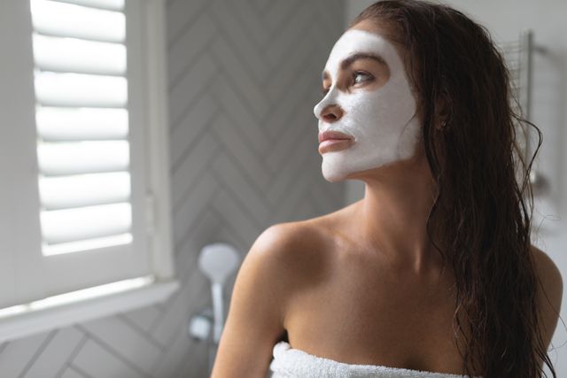 Woman with facial mask relaxing in bathroom, wrapped in towel, looking away. Ideal for content related to skincare routines, beauty tips, self-care practices, wellness blogs, and spa advertisements.