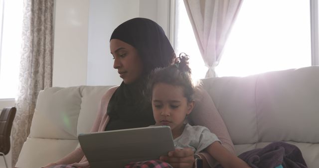 Front view of a young biracial woman wearing hijab with her young daughter in the sitting room, sitting on a sofa the girl using a tablet computer, the woman using laptop computer