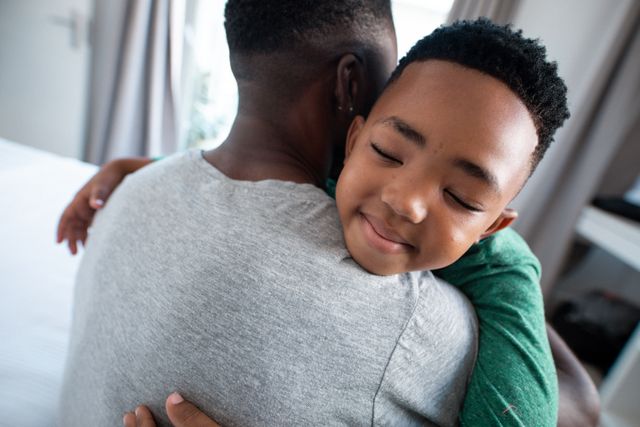 This image captures a tender moment between an African American father and his son hugging in a bedroom. The child is smiling with eyes closed, conveying a sense of happiness and security. This image is perfect for use in family-oriented content, parenting blogs, advertisements promoting family values, or any material highlighting the importance of father-child relationships.