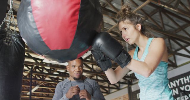 Female athlete boxing with a trainer in a gym. She is concentrating while hitting the punching bag, and the trainer is watching closely, providing guidance. Perfect for images depicting fitness training, workouts, and personal training sessions. Useful for fitness, sports, and health-related marketing, inspiration, and educational materials.
