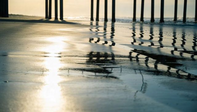 Sunlit pier pillars reflecting on wet sand at low tide with golden-hour glow