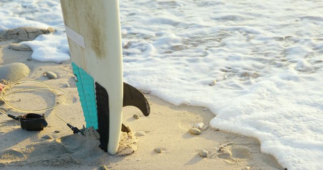 Surfboard partially buried in sand near ocean waves. Ideal for themes related to surfing, beach vacation, seaside activities, summer, and water sports.