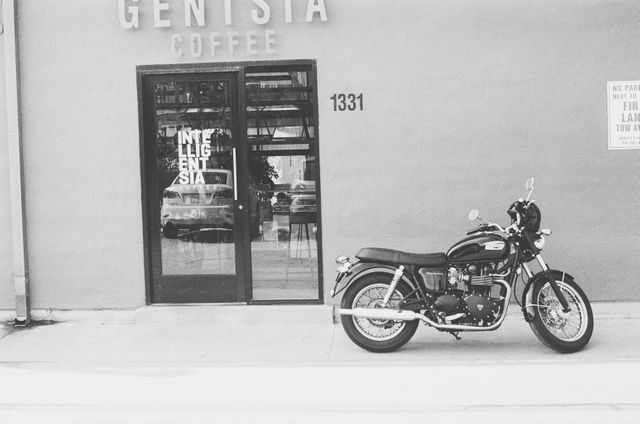 Vintage motorcycle parked in front of a coffee shop with large windows and door. Scene conveys a sense of nostalgia and retro urban lifestyle. Perfect for themes related to transportation, urban life, and minimalist aesthetics. Suitable for marketing campaigns targeting coffee shops, motorcycle enthusiasts, or stores with vintage and retro themes. Can also be used in web design for blogs or articles about city living or vintage motorcycles.