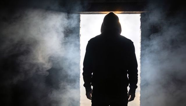 Hooded figure standing in backlit doorway with smoke, dramatic silhouette, suspense