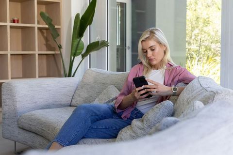 Young woman relaxing on gray sofa scrolling smartphone in bright minimalist living room