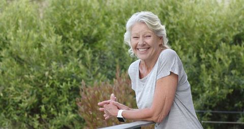 Senior woman leaning on balcony railing smiling while wearing smartwatch in garden