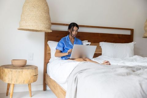 Woman working on laptop in blue silk pajamas on wooden-framed bed minimalist cozy bedroom