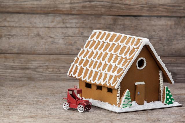 Gingerbread house decorated with icing and small toy car on rustic wooden background. Ideal for holiday greeting cards, festive advertisements, Christmas-themed blog posts, and seasonal social media content.