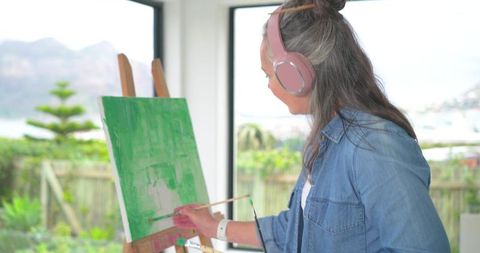 Mature woman painting green canvas listening with headphones in sunlit home studio