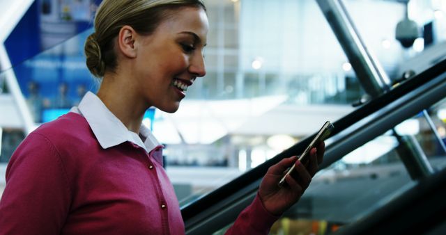 Businesswoman smiling while using smartphone on escalator in modern building. Ideal for content related to technology, communication, modern lifestyle, or business travel. Adds a touch of professional and contemporary ambience to articles or promotional materials.