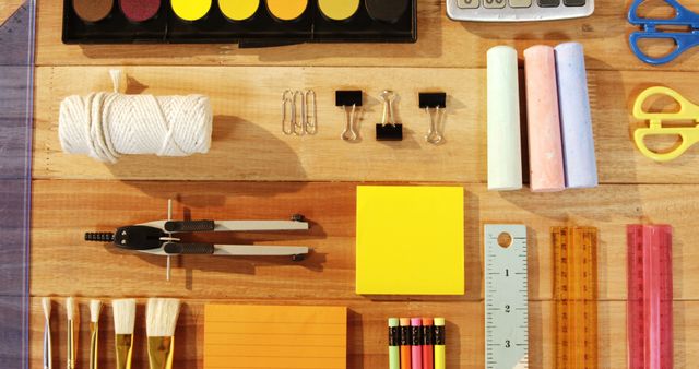 Flat lay of organized school and office supplies on a wooden desk. Includes various stationery items such as a set of paints, a calculator, scissors, brushes, pencils, rulers, notepads, paper clips, and binder clips. Ideal for illustrating themes of creativity, education, homework, preparation, or office work. Suitable for articles, blog posts, educational materials, or advertisement channels.