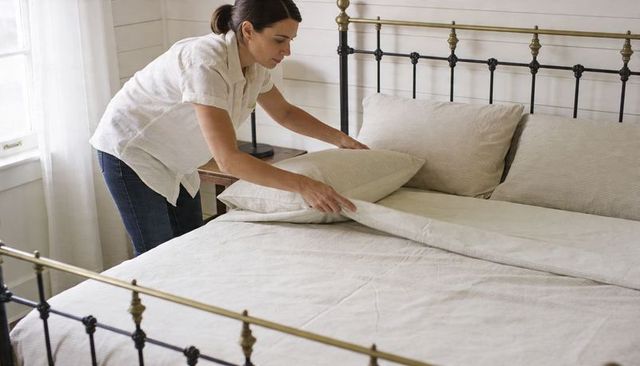 Making bed: middle-aged woman arranging linen duvet on brass bed frame in minimal bedroom
