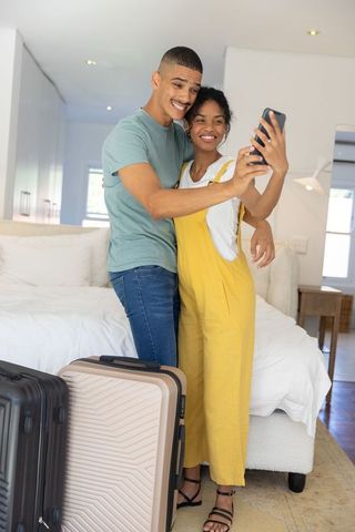 Young couple taking selfie with luggage in bright bedroom before trip