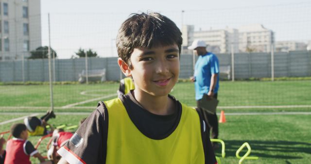 Young boy in sportswear smiling during outdoor soccer training session on a sunny day. Other children visible in background taking part in exercises, small hurdles can be seen. Soccer coach in blue shirt standing in the background with training equipment. Useful for promoting youth sports, outdoor activities, and fitness programs.