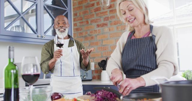 A joyful mature couple is cooking together in a home kitchen. The woman is chopping vegetables while the man is holding a glass of red wine and smiling. Use this image to depict concepts of a loving relationship, home cooking, culinary enjoyment, and happy domestic life.