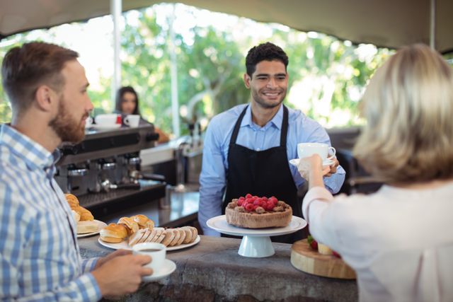 Waiter serving coffee to a customer at an outdoor cafe counter, with pastries and cake displayed. Ideal for use in hospitality, service industry, and coffee shop promotions. Highlights friendly customer service and casual dining atmosphere.
