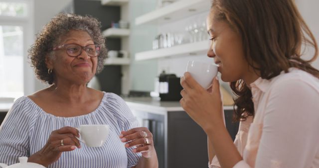 Happy senior african american woman and adult daughter sitting at table, drinking tea and talking. Lifestyle, free time, family and domestic life.