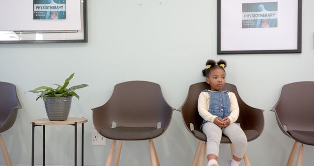 Young girl sits in a hospital waiting room on a modern chair, wearing casual clothes. Empty chairs and a potted plant are nearby. Suitable for use in healthcare advertisements, pediatric care campaigns, medical facility brochures, and articles about hospital experiences for young patients.