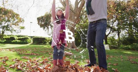 Father and daughter tossing autumn leaves while bitcoin hologram floating over sunlit park