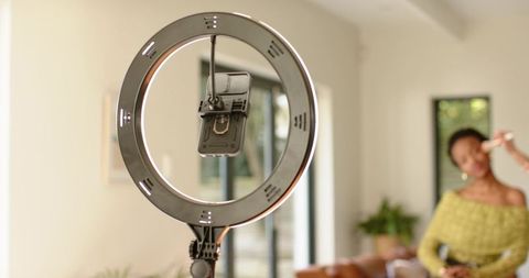 African american beauty creator applying makeup under ring light with phone clamp at home