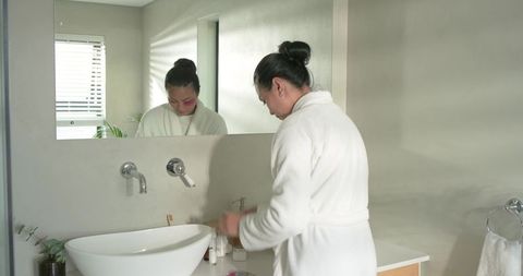 Asian woman performing morning skincare routine at modern bathroom vanity with white vessel sink