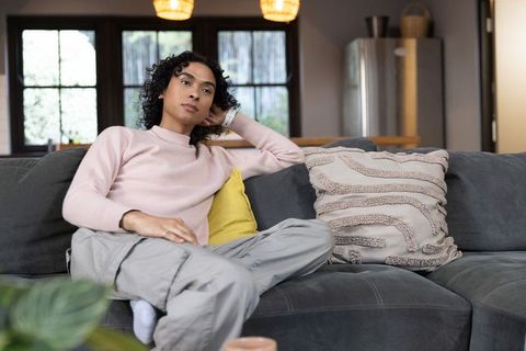 Non-binary person relaxing on modern gray sofa holding yellow pillow in cozy sunlit living room