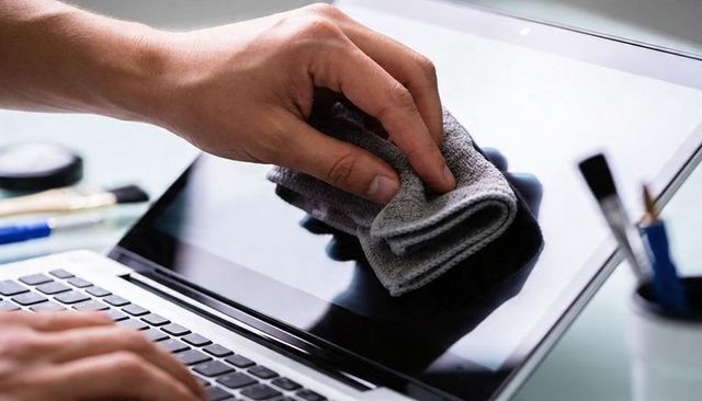 Hand wiping laptop screen with microfiber cloth near keyboard showing clear reflection