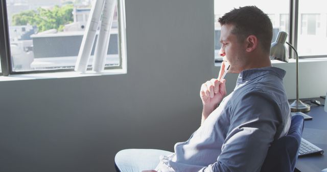 Thoughtful man sitting by window considering ideas. Ideal for business, productivity, and innovation-themed content. Useful for articles on workplace habits, mental health, and creativity.