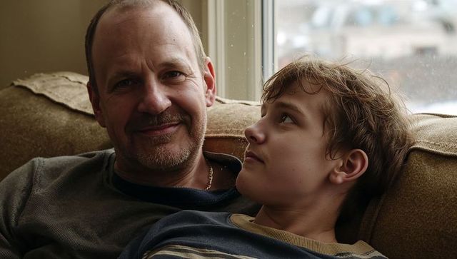 Smiling father holding teen son on cozy couch by window, sharing warm family moment