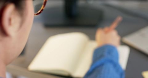 Woman writing in blank notebook at desk with glasses and laptop, workspace productivity concept