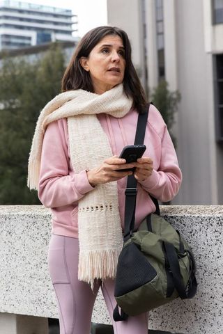 Woman checking phone while waiting outdoors in pink hoodie and cream scarf