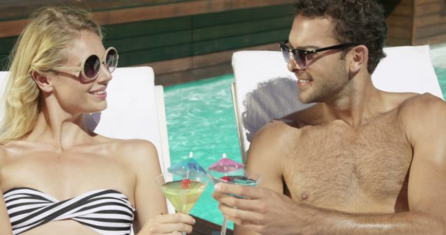 A man and woman are lounging poolside on sunbeds and toasting with refreshing cocktails. Both are wearing sunglasses and swimwear, basking in the sun. This can be used for travel promotions, leisure and relaxation marketing, summer vacation concepts, and luxury resort advertisements.