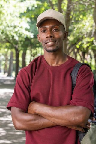 Confident adult man standing with arms crossed wearing cap and backpack in park