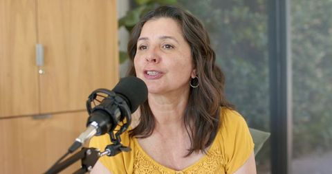 Mature woman speaking into mic recording podcast in home studio wearing yellow blouse