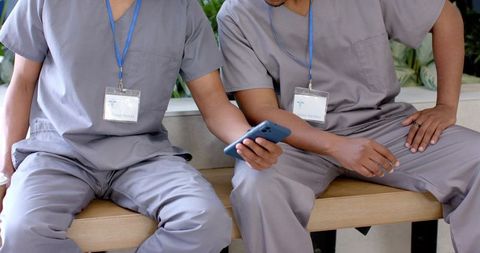 Two african american male healthcare workers sitting on bench sharing smartphone in scrubs